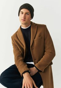 Brown wool blend coat with a tailored fit, single pocket, layered over a dark sweater. Model wears a ribbed dark brown knit beanie.