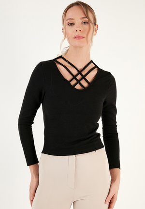 Black ribbed long-sleeve top featuring a V-shaped neckline with crisscross straps, fitted silhouette, and soft texture. Paired with light pants.
