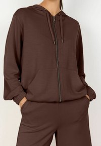 Bruine hoodie met rits, voorzien van voorzakken, elastische manchetten en een gladde textuur, gecombineerd met bijpassende broek.