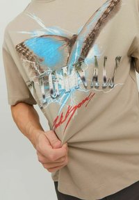 T-shirt in cotone beige con una grafica di farfalla blu, dettagli metallici e un design di testo audace con effetto invecchiato. Maniche corte.