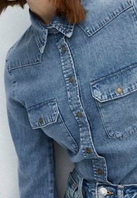 Chemise en denim bleu clair avec un col pointu, des manches longues et deux poches poitrine. Dotée de boutons métalliques et d'une texture légèrement délavée.