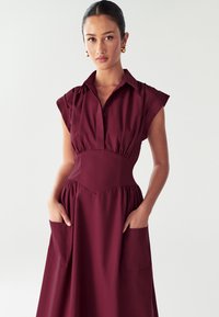 Abito camicia bordeaux con maniche corte, colletto a camicia, vita elasticizzata e due tasche frontali; realizzato in morbido tessuto texturizzato.