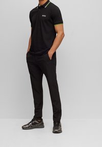 Svart pikétröja med tvåknäppning, gröna och vita krage detaljer, ihop med åtsittande svarta byxor och svarta sportskor.