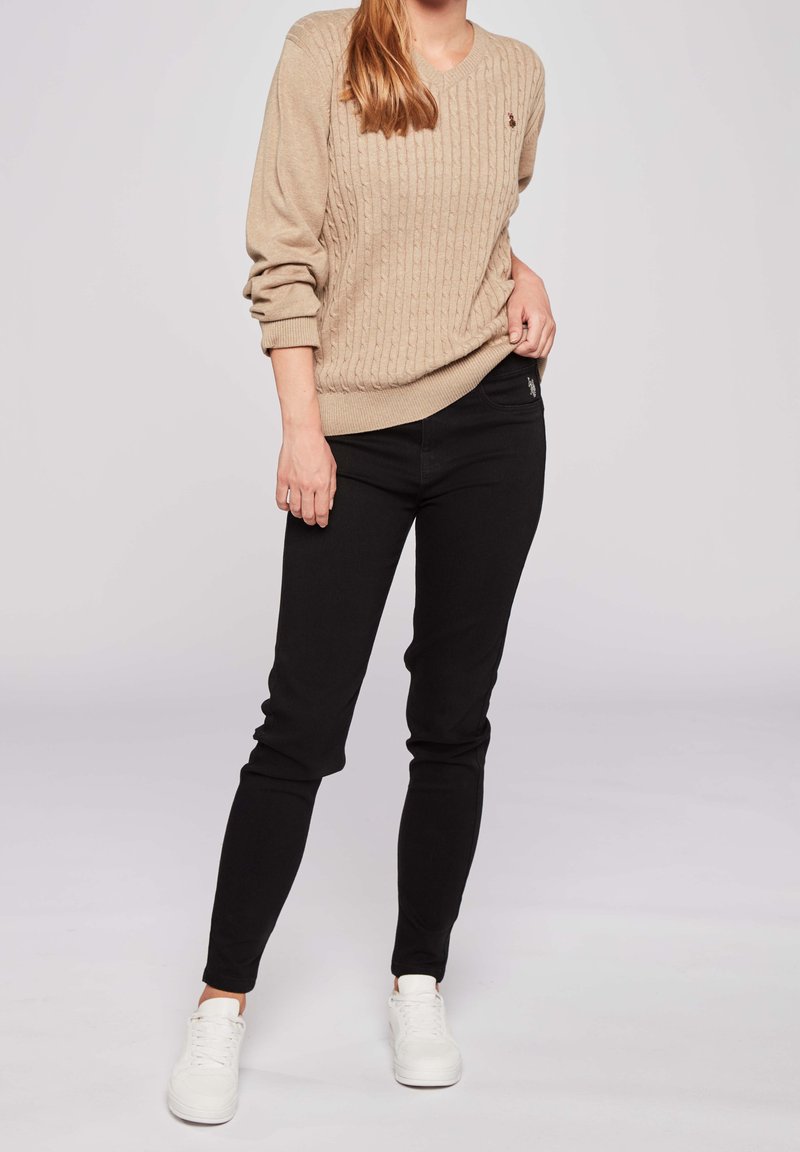 Jersey de punto en color beige con escote en V, combinado con jeans negros ajustados y zapatillas blancas. La prenda presenta un pequeño logo en el suéter.