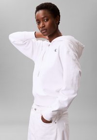 Sudadera blanca con capucha, hecha de algodón, que incluye un bolsillo frontal, cordón ajustable y un pequeño logo en el pecho. Se lleva con pantalones blancos, posando sobre un fondo gris.