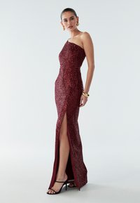 Burgundy sequined evening gown with a one-shoulder design, side slit, and fitted silhouette. Accented by a smooth, glossy texture.