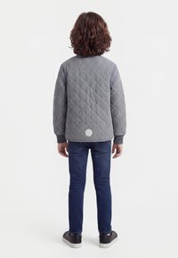 Barn med krøllet hår set bagfra, iført en grå quiltet jakke, mørkeblå jeans og sorte sneakers med hvide såler.