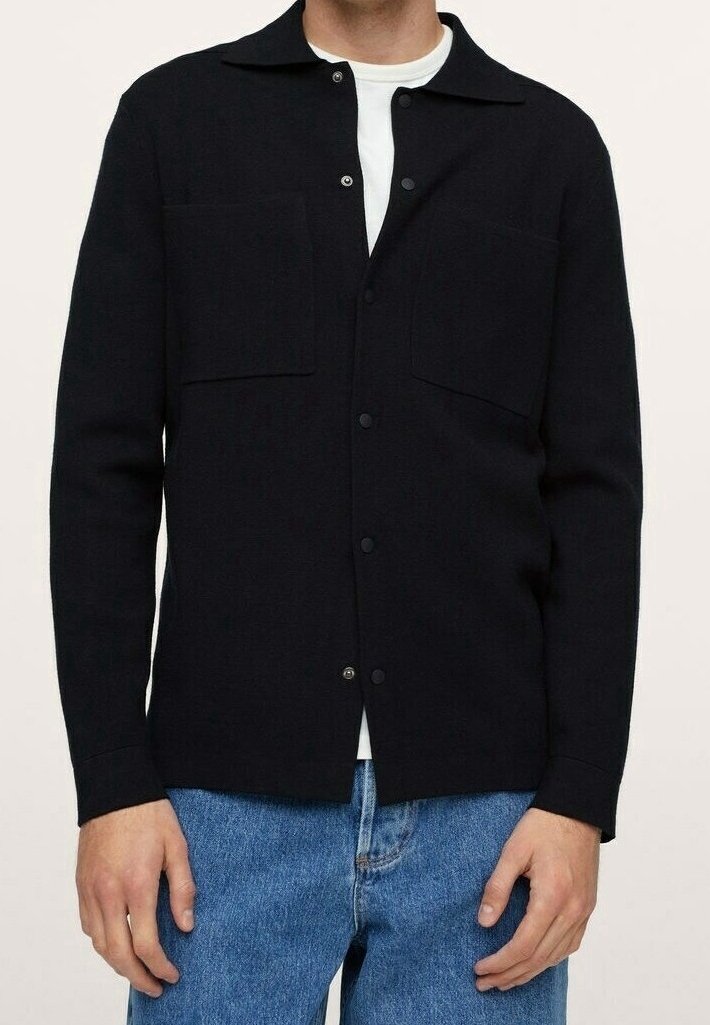 Homme portant une veste noire boutonnée avec deux poches poitrine sur une chemise blanche et un jean bleu, debout avec les mains détendues sur les côtés.