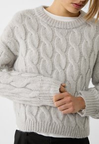 Hellgrauer Kabelstrickpullover mit Rundhalsausschnitt, gerippten Bündchen und strukturierten Mustern. Aus weichem, flauschigem Material gefertigt.