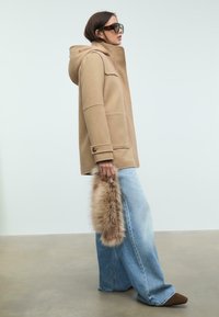 Cappotto di lana beige con cappuccio, dotato di ampie tasche e dettagli con bottoni, abbinato a jeans a gamba larga di un blu chiaro e una finitura in pelliccia sintetica.