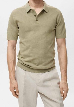 Hombre que lleva una camisa polo beige de manga corta con tres botones, combinada con pantalones beige claro, de pie frente a un fondo sencillo.