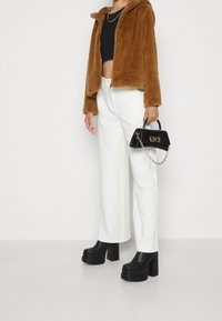 Chaqueta con cremallera de piel sintética marrón, top corto negro, pantalones anchos blancos y botines negros con plataforma. Pequeño bolso negro con cadena.