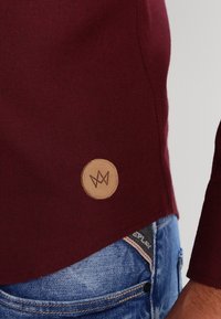Camicia in tessuto bordeaux con un'etichetta rotonda in pelle beige che presenta un logo, abbinata a jeans in denim blu con un'etichetta vicino alla vita.