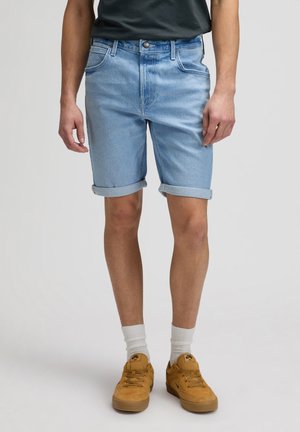 Uomo che indossa pantaloncini di jeans azzurri risvoltati, calzini bianchi e scarpe da ginnastica color cammello, in piedi con le mani rilassate ai lati.