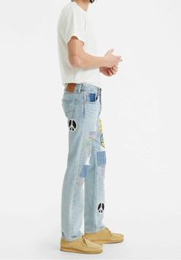 Pantalones de mezclilla azul claro con parches y apliques coloridos, que presentan un símbolo de paz, combinados con una camiseta blanca y zapatos beige.