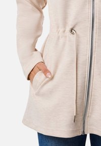 Veste beige clair avec un motif texturé, fermeture éclair à l'avant, taille à cordon de serrage et poches latérales. Comprend des manches longues et une coupe décontractée.