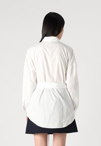 Camicia bianca a maniche lunghe con vita legata, caratterizzata da un colletto a punta e orlo curvo, abbinata a una gonna blu navy scura. Tessuto dalla trama liscia.