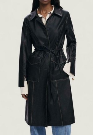 Kvinde iført en lang sort læder trench coat med knaplukning, taljebånd, klaplommer, over en lys skjorte og blå jeans.