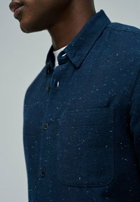 Camisa de botonadura en azul marino hecha de tela texturizada, con un bolsillo en el pecho y un sutil patrón moteado. Cuello redondeado.