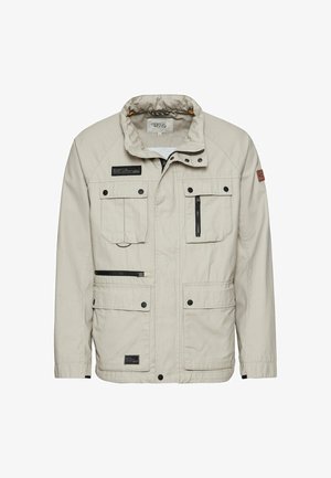 Veste beige à poches multiples avec col montant, fermeture éclair à l'avant et quincaillerie contrastante. Fabriquée en tissu léger avec une texture lisse.