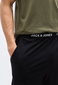 Άτομο που φοράει μαύρο παντελόνι με ζώνη μέσης με την επωνυμία "JACK & JONES" και ένα πουκάμισο χακί πράσινο, με το χέρι στην τσέπη.