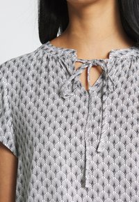 White blouse with black geometric pattern, short sleeves, keyhole neckline tied with a bow, soft fabric with a smooth texture.