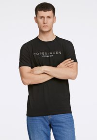 Młody mężczyzna z krótkimi włosami, złożonymi ramionami, ubrany w czarną koszulkę z napisem "Copenhagen Lindbergh" i niebieskie dżinsy, stojący na tle białym.