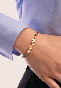 Pulsera de oro con "mama" en cursiva, adornada con cuentas coloridas en tonos de rosa y negro, mostrada en una muñeca con una camisa azul.