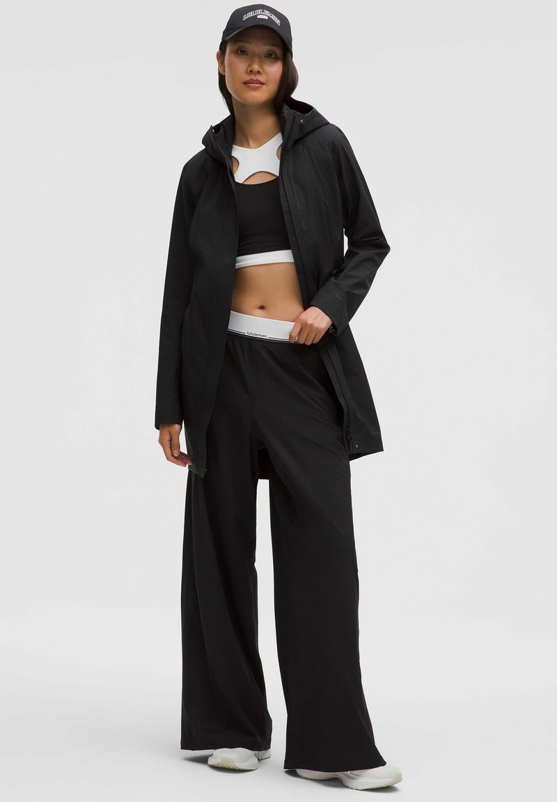 Mujer vistiendo pantalones anchos negros, una chaqueta con capucha negra, un top corto en blanco y negro, zapatillas blancas y una gorra negra con texto blanco.