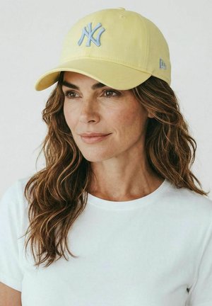Mujer con cabello castaño ondulado, que lleva una gorra amarilla clara de los New York Yankees y una camiseta blanca lisa, mirando ligeramente hacia un lado sobre un fondo blanco.