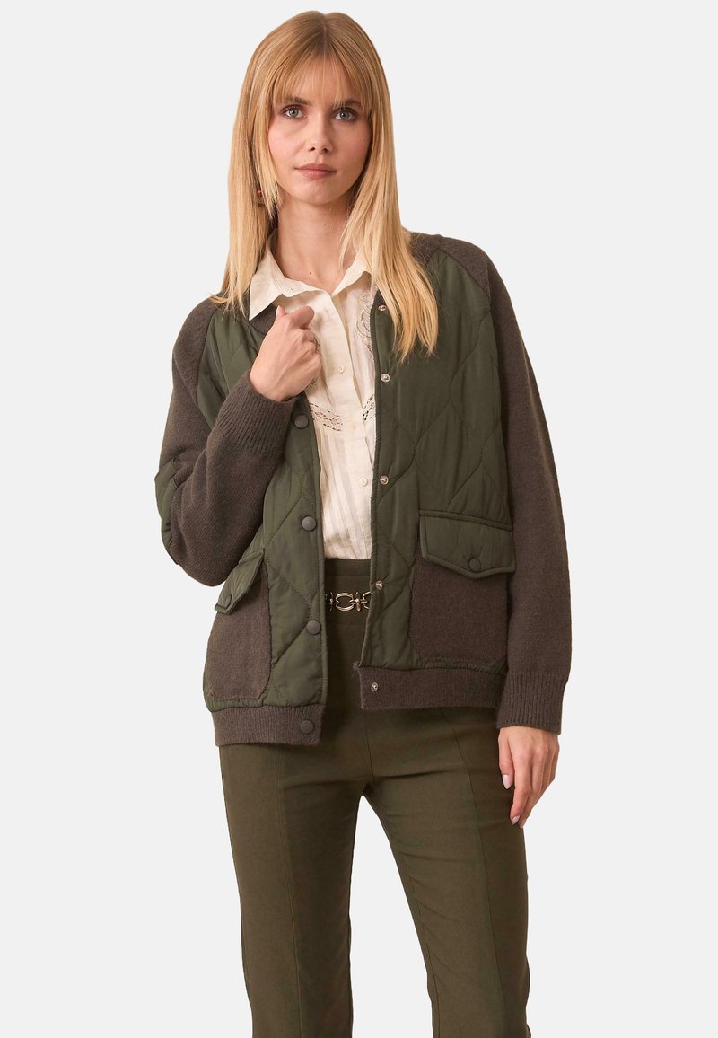 Mujer con cabello rubio liso viste una chaqueta acolchada verde sobre una blusa blanca y pantalones verde oliva, posando frente a un fondo liso.