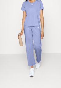 Person i lila Nike-träningskläder med vita sneakers som håller en beige vattenflaska, går mot en enkel ljus bakgrund.