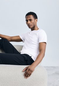 Fehér pólóban és fekete nadrágban ülő férfi egy világos textúrájú kanapén, egy sima világosszürke háttér előtt.