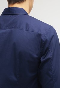 Person som bär en mörkblå skjorta med krage, visad från baksidan, med synliga sömnadsdetaljer på axlarna och ärmarna.