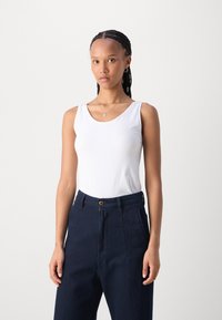 White sleeveless tank top made of soft fabric, paired with high-waisted navy blue textured trousers featuring front pleats and a button accent.