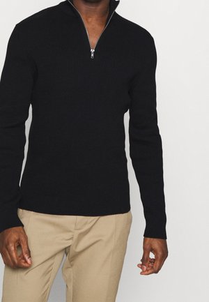 Mężczyzna w czarnym prążkowanym swetrze z krótkim zamkiem przy kołnierzu i beżowych spodniach na jednolitym tle.