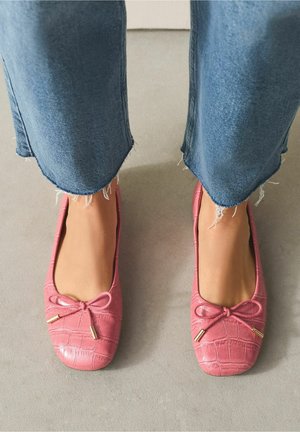 Pieds portant des ballerines roses texturées avec petits nœuds et bouts dorés, associées à un jean bleu à ourlet effiloché sur un sol gris.