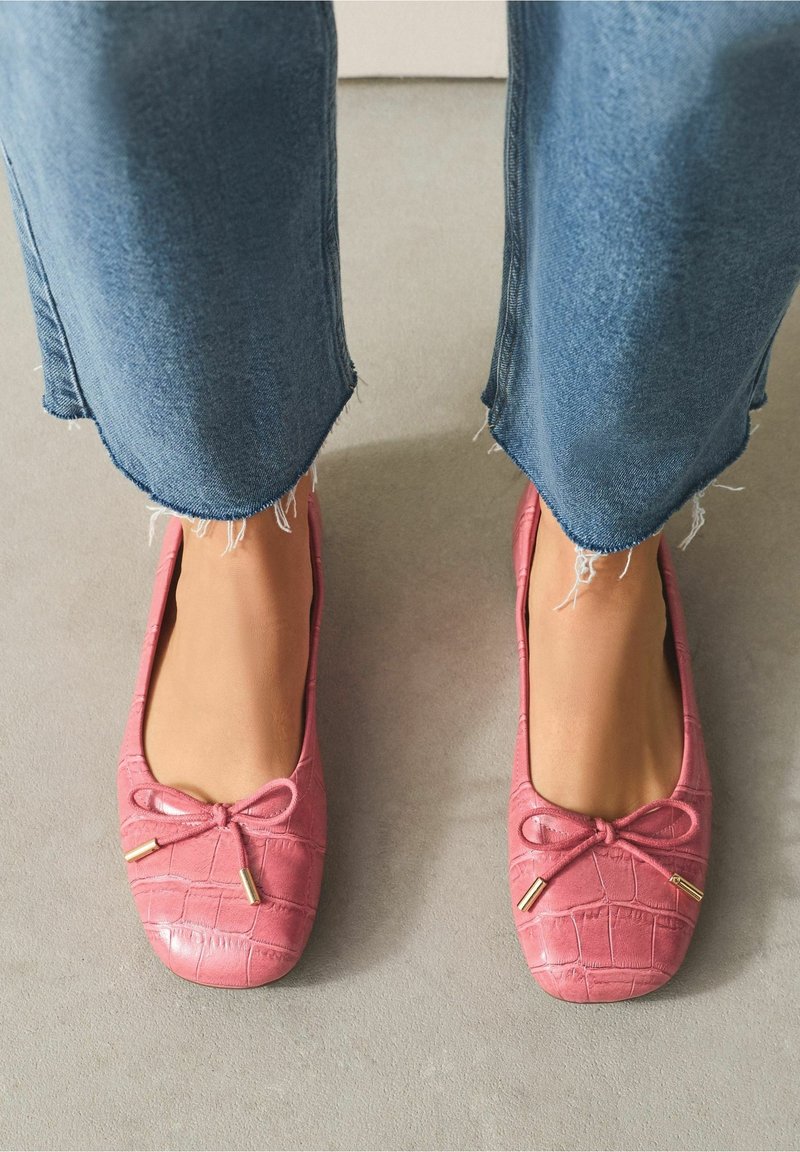 Pieds portant des ballerines roses texturées avec petits nœuds et bouts dorés, associées à un jean bleu à ourlet effiloché sur un sol gris.