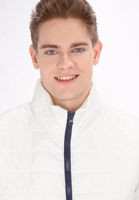 Weißer Pufferjacke mit hohem Kragen und einem marineblauen Reißverschluss. Der Stoff hat eine gesteppten Textur mit horizontalen Nähten.