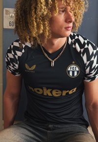 Schwarzes Sporttrikot mit einem Chevron-Muster auf den Ärmeln, goldene Schriftzüge, Logo auf der Brust und einem runden Ausschnitt, kombiniert mit hellen Jeans.