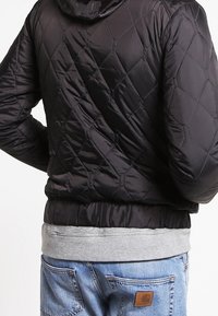 Svart vadderad jacka med huva, med ett texturerat mönster och ribbad grå fåll. Bärs över ljusblå denimbyxor med en logotyppatch.