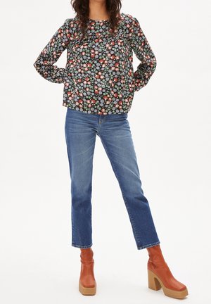 Kvinde iført blomstret langærmet top, lige blå jeans og brune platform-ankelstøvler står med hænderne bag ryggen.