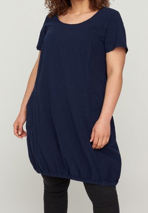Vrouw draagt een losse marineblauwe korte mouw jurk met zijzakken en een geplooide zoom over zwarte leggings.
