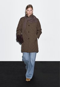 Hnědý prošívaný kabát s černými knoflíky, kombinovaný se světle modrými širokými džíny. Šedý šátek je omotán kolem krku a v rukou jsou tmavě hnědé rukavice.