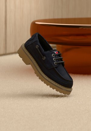 Marineblaue Veloursleder-Schnürschuhe mit dicker beiger Gummisohle, schwebend über einem beigen Teppich mit braunen Möbeln im Hintergrund.