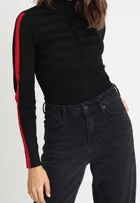 Suéter de punto negro de cuello alto con textura de rayas verticales, que presenta acentos rojos en las mangas. Combinado con jeans negros de tiro alto.