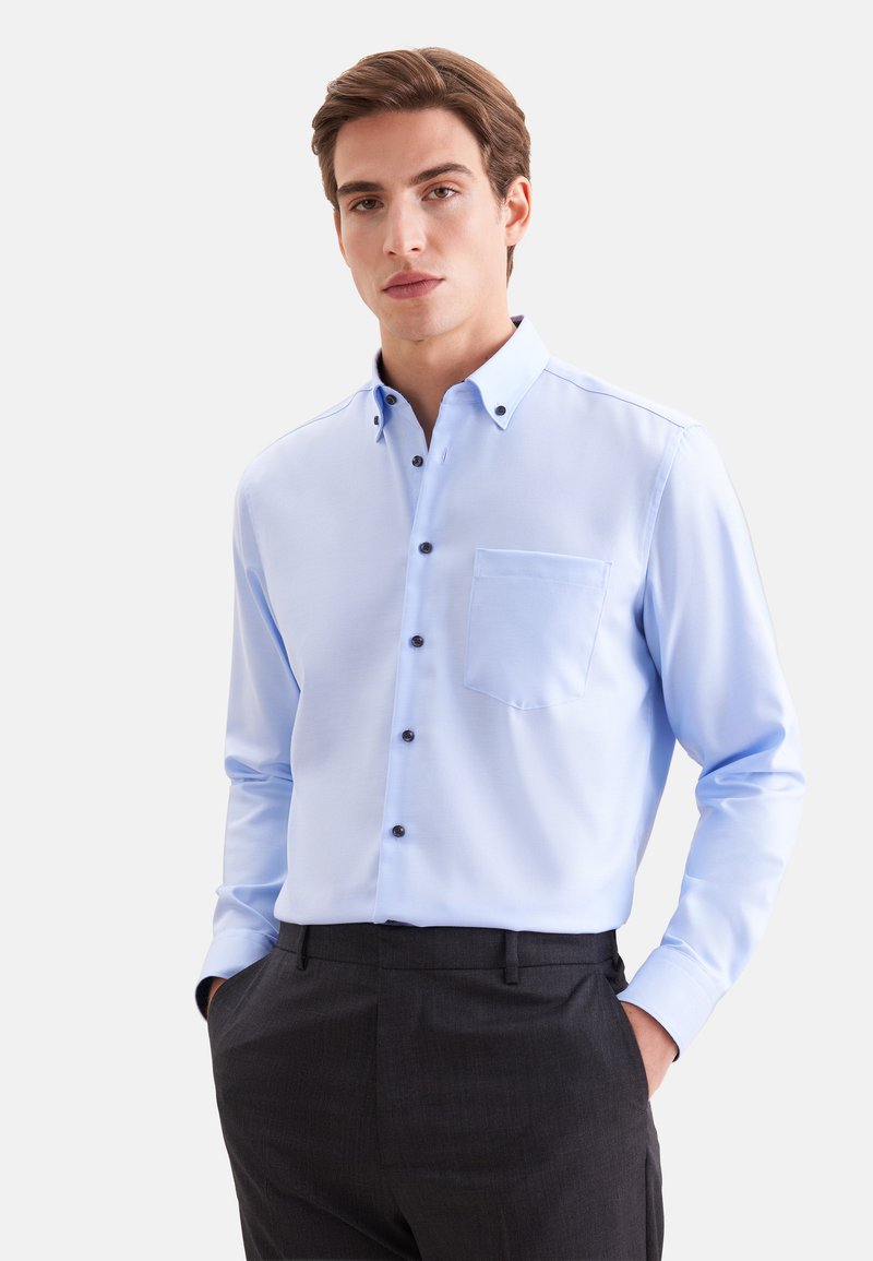 Jeune homme portant une chemise bleu clair boutonnée avec une poche poitrine et un pantalon gris foncé, debout les mains dans les poches.