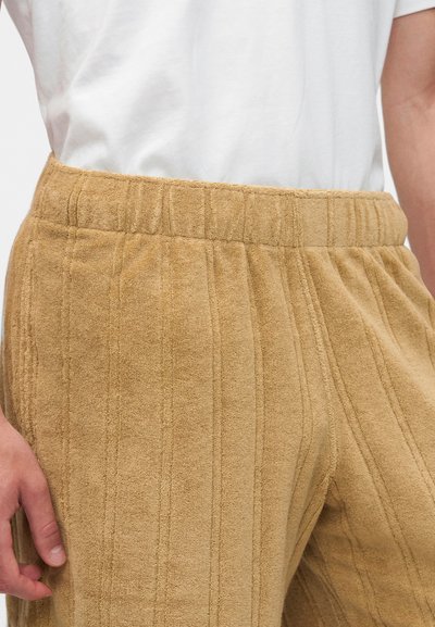 Pantalon lounge beige texturé avec rayures verticales et ceinture élastique porté avec une chemise blanche, main reposant sur le côté.