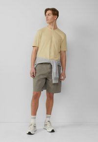 Beige katoenen t-shirt, grijze sweatshirt om de taille gebonden, olijfkleurige shorts, witte sneakers met zwarte accenten en gestreepte sokken. Eenvoudige ontwerpelementen.