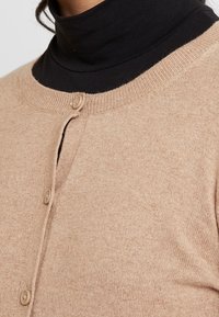 Beige kofta i mjukt stickat tyg, med rund halsringning, två knappar och ribbstickade detaljer vid kragen och knappslån.